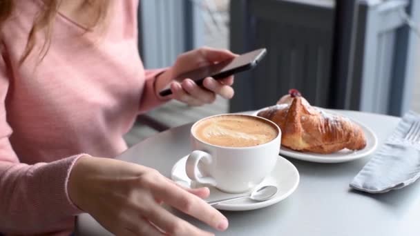 坐在咖啡馆桌旁的女人 一边喝咖啡 一边用智能手机 慢镜头 女孩在餐厅平台上吃午饭的时候 在智能手机上滚动她的新闻 关门了 — 图库视频影像