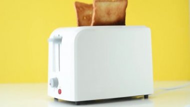 Roasted toast bread popping up from white toaster on yellow background, hand takes out toasts from toaster