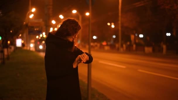 女性は彼女の時計を見て 夜の街で携帯電話で誰かを呼び出します焦点を当てたぼやけた街路灯と — ストック動画