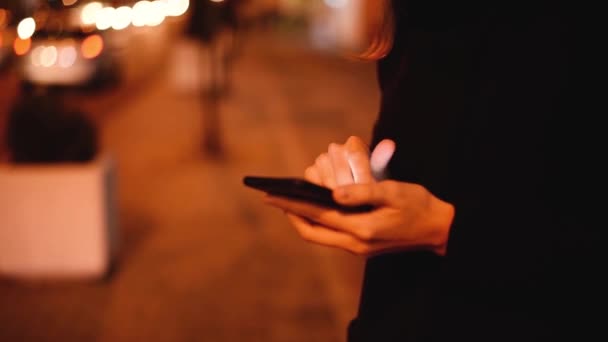 Femme utilisant un téléphone portable dans la ville de nuit. Gros plan des mains féminines tenant le smartphone contre les lampadaires déconcentrés 
