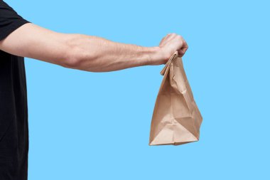 Yemek teslimatı konsepti. Siyah üniformalı bir kurye mavi arka planda yiyecek taşıyan bir kese kağıdı tutuyor.