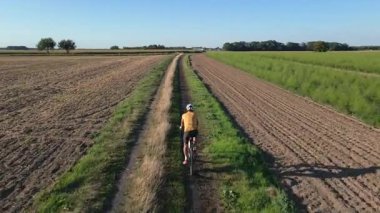 Man wearing helmet and sportswear riding gravel bike on dirt road between farmland and green fields at sunset. Cyclist training on bicycle. Active lifestyle concept