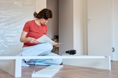 Mobilya montajı sırasında yerde oturmuş kullanma kılavuzunu okuyan bir kadın. Yerde aletlerin ve yeni yatağın parçalarının yanında oturan biri. DIY ev geliştirme ve mobilya kurulumu kavramı