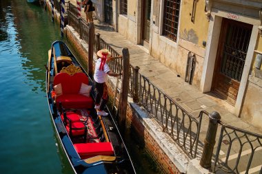 Gondolier standing on gondola docked by canal in Venice, Italy. Venetian gondola with red seats and ornate decorations. Concept of travel and tourism