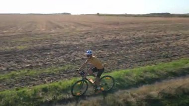 Man wearing helmet and sportswear riding gravel bike on dirt road between farmland and green fields at sunset. Cyclist training on bicycle. Active lifestyle concept