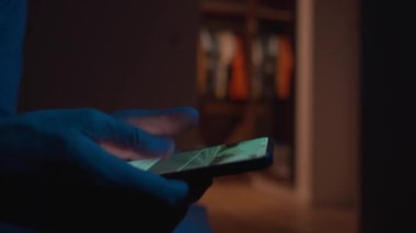 Man using smartphone at night. Close up of hand holding smartphone, swiping and scrolling social media feed on glowing screen in dark room