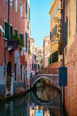 Venetian narrow canal with small stone bridge and colorful historic building facades with open shutters. Concept of travel, sightseeing and European cultural heritage.