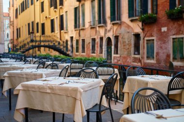 Empty outdoor restaurant tables with tablecloths and cutlery in Venice, Italy. Street cafe with bridge over Venetian canal on background in morning. Concept of dining, tourism and travel