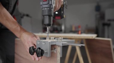 Close-up of carpenter drilling hole in wooden panel using drill and metal jig tool for precision. Man working in home workshop. Concept of woodworking, furniture making and DIY home projects