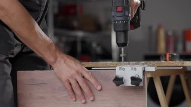 Close-up of carpenter drilling hole in wooden panel using drill and metal jig tool for precision. Man working in home workshop. Concept of woodworking, furniture making and DIY home projects
