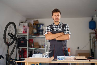Portrait of carpenter in checkered shirt and overalls standing with crossed arms in home workshop surrounded by tools and wooden materials. Concept of woodworking and professional craftsmanship