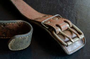Çifte metal tokalı eski bir deri kemer çatlamış. Karanlık bir masaya geniş, kirli bir kemer ya da tasma yakın plan. Koleksiyonluk eski moda aksesuarlar. Seçici odak.