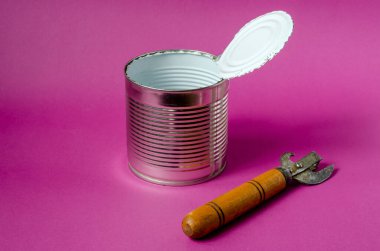 An old can opener lies next to an empty tin can on a purple background. Open tin can with white food enamel inside. Hiking, travel. Close-up. Copy space. clipart
