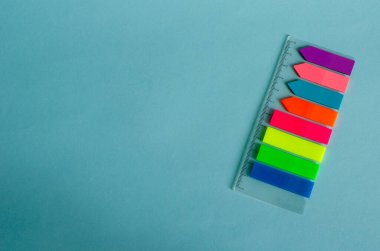 Açık mavi bir yüzey üzerinde yatay olarak bir tablo üzerinde bir dizi renkli yer imi bulunur. Cetvel ve çizgili çıkartmalar. Düz yatıyordu. Leylak, mercan, mavi, turuncu, kırmızı, sarı, yeşil, mavi. Boşluğu kopyala.