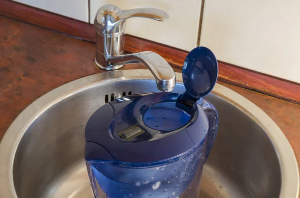 Water filter jug in the kitchen sink. Plastic jug filled with running water with blue lid. Top view at an angle.