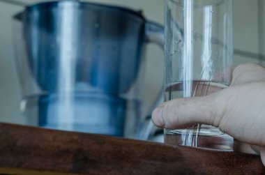 Mutfak masasında bir bardak içme suyu olan orta yaşlı beyaz bir adamın eli. Arka planda su testisi var. Su iç. Yakın plan. Seçici odak.