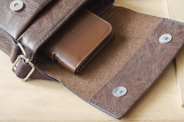 Brown leather wallet inside small shoulder bag. An open bag and brown leather wallet on wooden background. Top view at an angle. Selective focus.