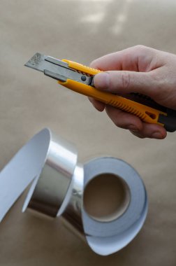Orta yaşlı beyaz bir adam elinde turuncu bir bıçak tutuyor. Arka planda folyo bandı rulosu. Isı ve nem yalıtım hizmetleri. Modern yalıtım malzemeleri. Seçici odak.