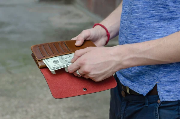 The hands of a young man of European ethnicity remove a five-dollar bill from a purse. Red braided charm bracelet and silver rings on the man\'s fingers. Lifestyle. Selective focus.