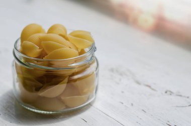 Beyaz ahşap arka planda cam kavanozda çiğ Conchiglie Pasta. Pişmemiş makarna servisine yakın çekim. Yiyecek depolamak için çevre dostu ambalaj. Bir açıdan yan görüş. Seçici odak.