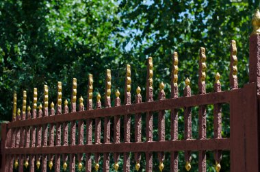 Yeşil ağaçların arka planında eski metal çitler. Kırık boyalı ve yaldızlı kaplamalı kırmızı bir çit. Turizm ve seyahat. Yan görüş. Seçici odak.