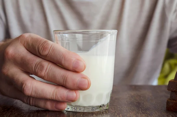 Close-up of man\'s hand with half-empty glass of kefir. Hand holds fermented milk drink on wooden table. Drink kefir. Adult male, Caucasian. Age 44-45. Lifestyle.
