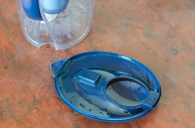 Mutfak masasındaki suyu temizlemek için bir sürahinin üst kapağını kapat. Sürahiye yerleştirilmeden önce ıslak mavi kapak. Eski bir su filtresini yenisiyle değiştirme süreci. Bir serinin parçası.