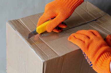 Koruyucu eldiven giyen bir işçi bir karton kutuyu açar. Bıçaklı bir el paketin yapışkan bandını keser. Lojistik ve teslimat servisi. Seçici odak.