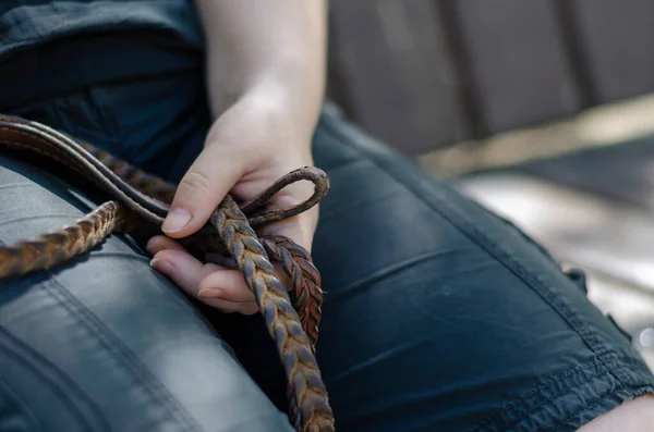 Close-up of a woman\'s hand with a leather leash. An adult woman holds a wicker leather leash for a pet sitting on a park bench. Rest during a walk. An active lifestyle.