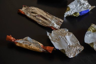 Siyah bir masada rastgele boş şeker paketleri. Kırıntıları ve lekeleri olan çok renkli folyo kağıtları. Çikolatalı şekerler. Şekerleme. Seçici odak.