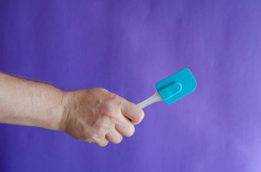 Mutfak silikon spatula elinde. Bir erkek el mavi arka planda plastik saplı bir mutfak spatulasını tutuyor. Seçici odak.