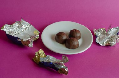 Pembe arka planda beyaz bir tabakta çikolatalı şekerler. Buruşuk şeker ambalajları arasında bir porsiyon hamur işi. Tatlı ikramlar. Seçici odak.