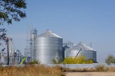 Mavi arka planda modern bir tahıl terminali inşa ediliyor. Metal silindirli silo ve yükleme için metal yapıyı destekleyin. Tarım işi. Tahıl ihracatı.