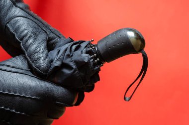 Siyah deri eldivenli eller mercan zemin üzerinde şemsiye tutuyor. Yağmur ve rüzgar koruma aksesuarı iki elle tutulur. Bir açıyla üstteki görünüm.