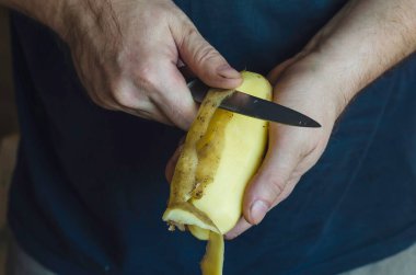 Olgun bir adam mutfak bıçağıyla çiğ patates soyar. Kafkasyalı bir adamın ellerinin yakın çekimi. Yemek hazırlama süreci. 44 yaşındasınız. Seçici odak.