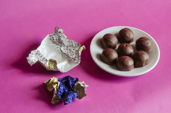 Chocolate candies in a white saucer on a pink background. A serving of Pastry among the crumpled random candy wrappers. Sweet treats. Selective focus.