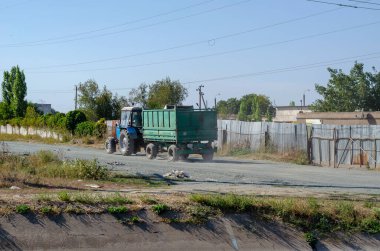 Nikolaev, Ukrayna - 15 Eylül 2020: Mavi bir traktör tozlu kırsal bir yol boyunca yeşil bir römorku çekiyor. Tarım mühendisliği.