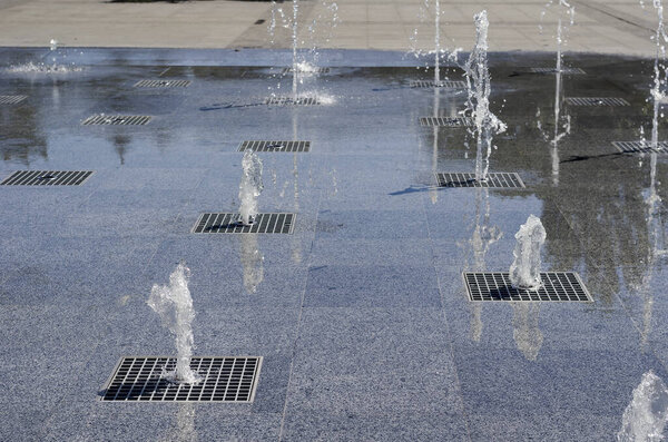 Water jets of the fountain in the city square. Jets of cool clear water gushing from the ground. Hot summer day.