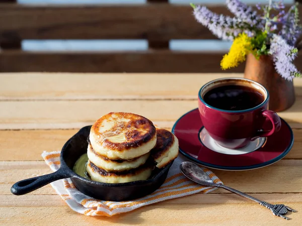 Cheesecakes are located on a small frying pan with an espresso cup. In the background is a bouquet of flowers. Located on a wooden brown board. Partial focus. — Stock Photo, Image