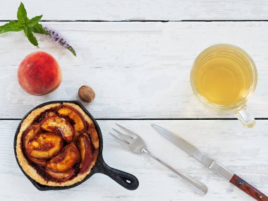 Beyaz ahşap vintage arka plan üzerinde yaz şeftali pasta. Kekin yanında çay, şeftali ve nane yaprakları olan bir fincan vardır. Çatal bıçak, bıçak ve çatal. Üst te. Metin için boş alan.