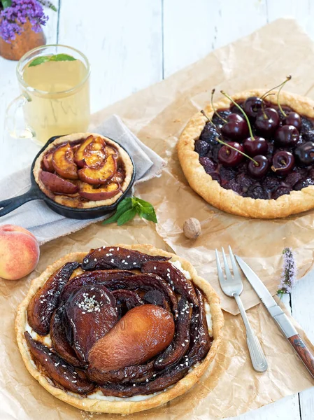 Summer cakes made from cherries, pears and peaches. Pies are located on a white wooden vintage background. In the background is a cup of tea, mint leaves and a bouquet of wildflowers. Close-up. — Stock Photo, Image