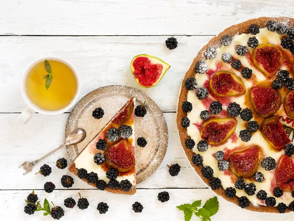 A piece of homemade pie with figs and blackberries is located on a vintage plate. Nearby is a pie and a cup of green tea with mint. Wooden vintage background. Next to the pie are blackberries, cut fig