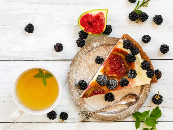 A piece of homemade pie with figs and blackberries is located on a vintage plate. Nearby is a cup of green tea with mint. Wooden vintage background. Next to the pie are blackberries, cut figs and cutl