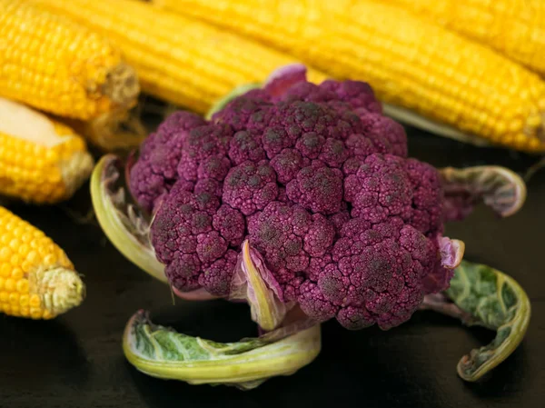 Verse, rijpe oren van maïs en vorken van paarse bloemkool. Gelegen op een zwart houten oppervlak. Close-up. — Stockfoto