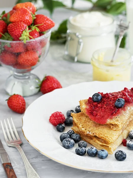 Pancakes with jam are located on a white plate, around the pancakes on a plate are fresh berries. Near jam and a cup of sour cream. Breakfast, festive food, christmas. Gray background. Close-up. The t — Stock Photo, Image