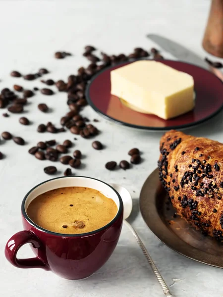 Burgundy color coffee cup with espresso and croissant. In the background are fresh coffee beans, butter and cezve. Located on a gray background. Close-up.