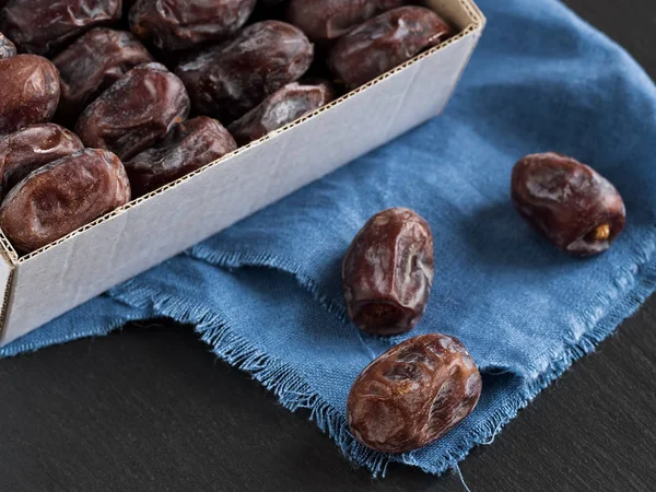 Oriental dates located in a box closeup. Nearby on a blue napkin are three dates. Black stone background. Selective focus, low depth of field, bokeh. — Stock Photo, Image