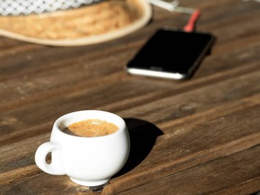 Ahşap bir masada kokulu espresso kahve. Arka planda bir hasır şapka ve bir telefon var. Close.