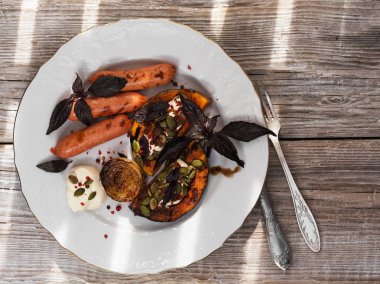 Beyaz seramik tabakta ızgara balkabağı, soğan ve sosis parçaları. Kremalı ve balzamik soslu. Balkabağı tohumu ve fesleğenle süslenmiş. Ahşap vintage yüzey. Çatal bıçak üst görünümü. Yatay yerleşim planı. P
