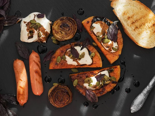 Grilled slices of pumpkin onions, bread and sausages on a stone board. Cream sauce and balsamic. Decorated with pumpkin seeds and basil. Stripes from sunshine. Top view.